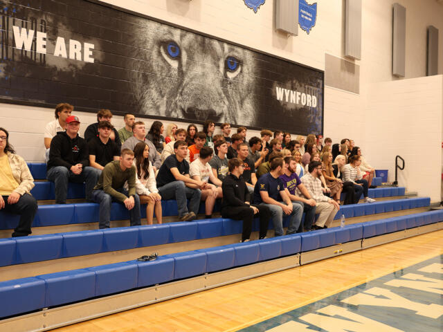 A large group of students sitting on blue gym bleachers in a school gym, posing for a group photo under a wall mural that reads 'WE ARE WYND' and features a lion's eyes close-up.