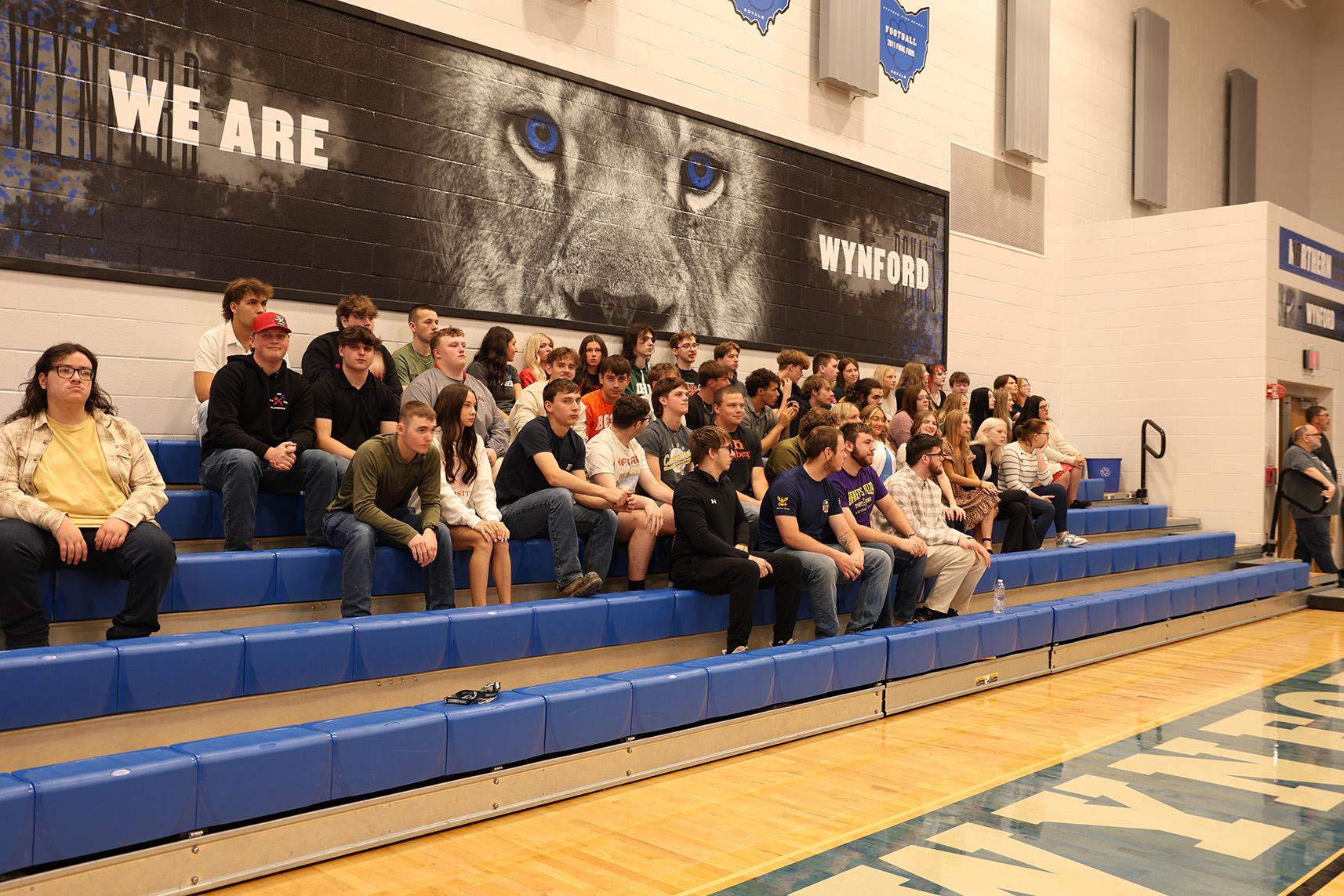 A large group of students sitting on blue gym bleachers in a school gym, posing for a group photo under a wall mural that reads 'WE ARE WYND' and features a lion's eyes close-up.