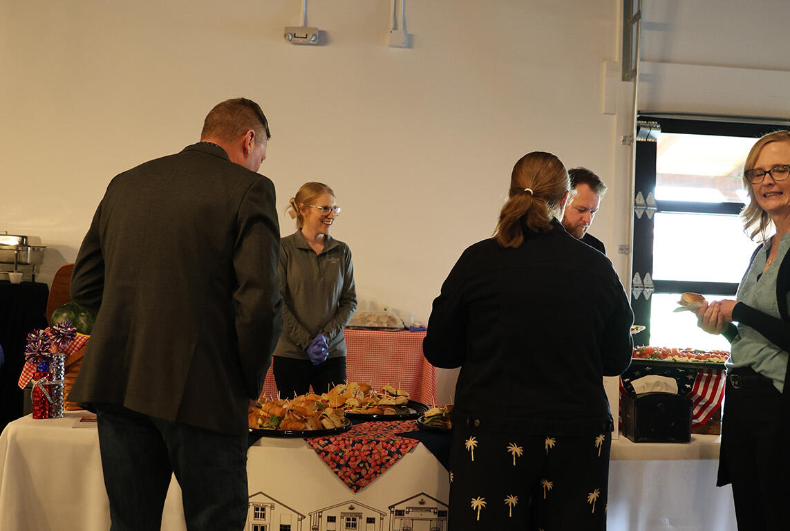 People stand at a buffet table indoors, selecting food from trays as others chat nearby; a banner with 'The Pickwick Place' is visible on the front of the table.