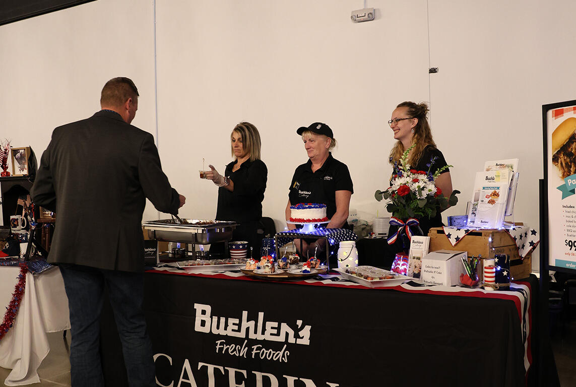 Catering booth at an indoor event with a long table covered in a black Buehlen Fresh Foods Catering banner, staff behind the table, a man in a blazer approaching the food, and a cake on display in the center.