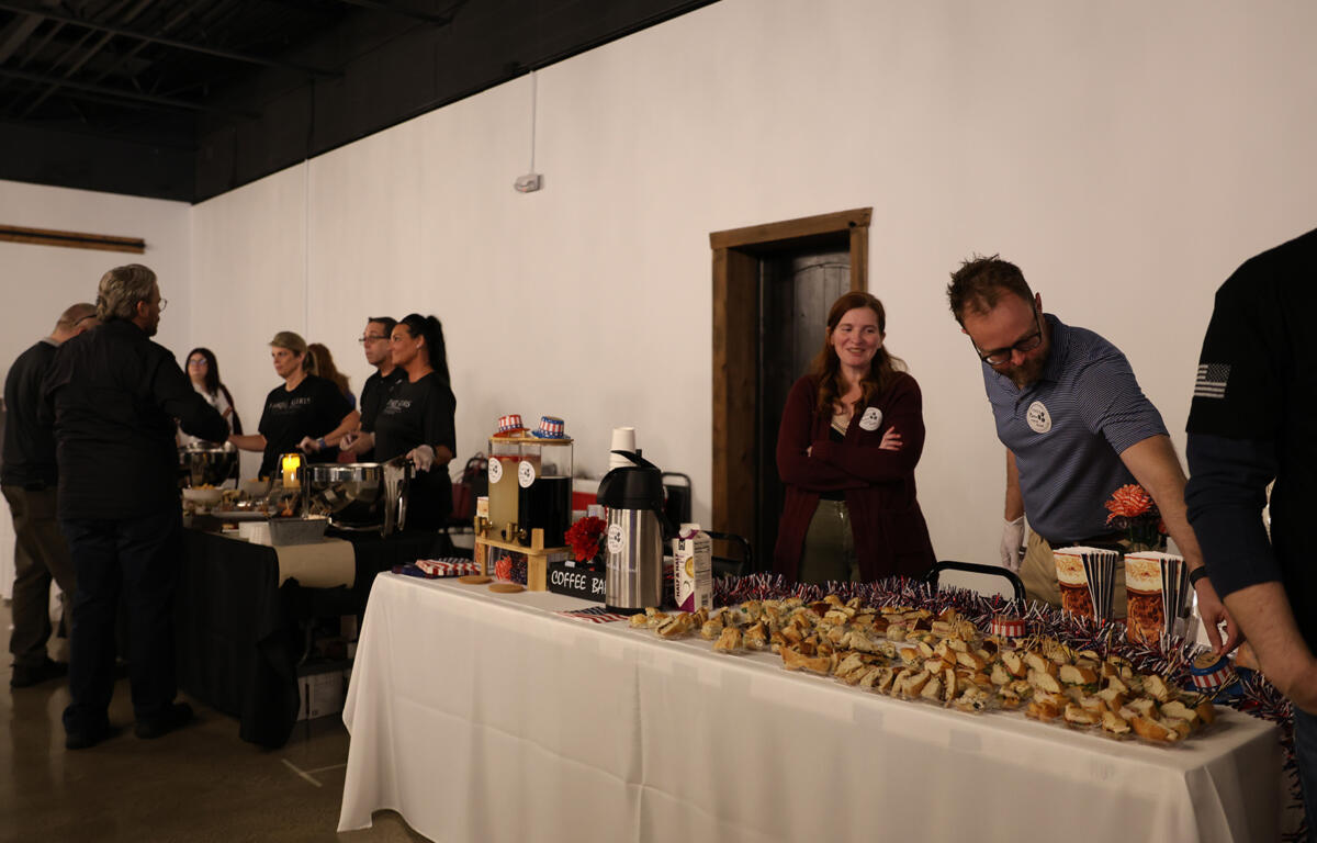 People gather around a long buffet table at an indoor event, with volunteers serving food and coffee on white tablecloths.