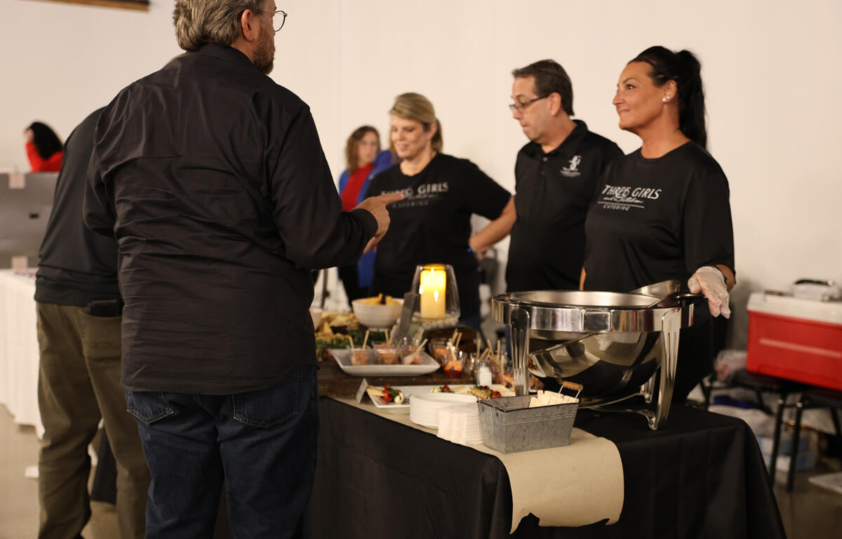Catering staff in black shirts behind a buffet table with chafing dish and bite-sized appetizers for guests in a banquet setting