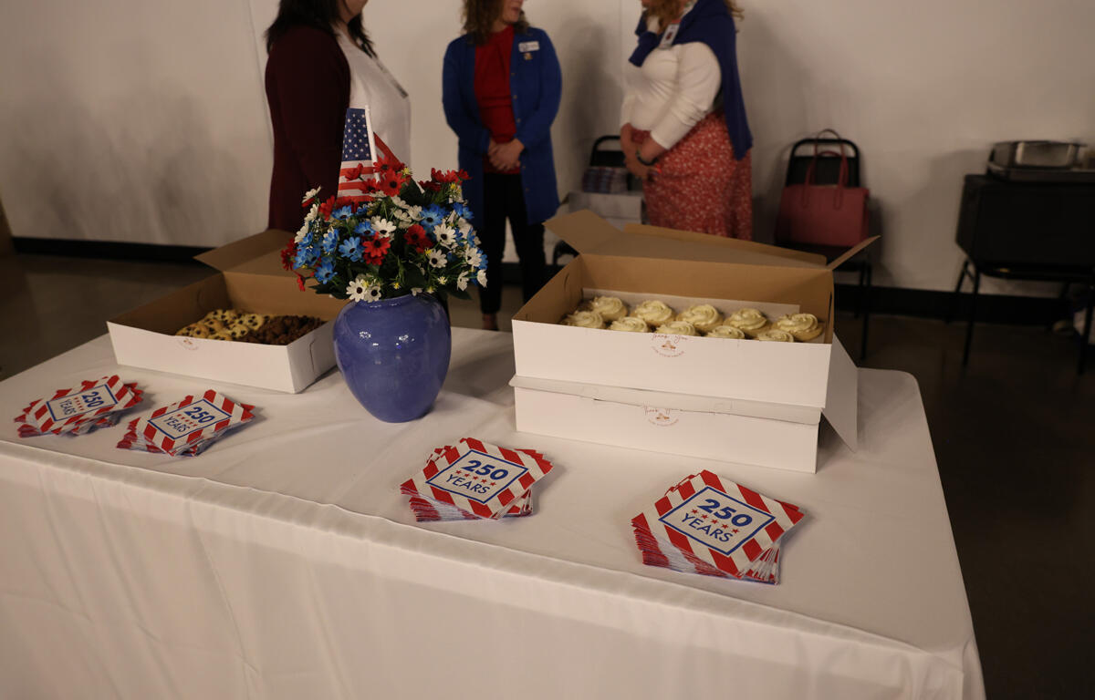 Blue vase of red, white, and blue flowers with a small American flag, flanked by two open boxes of cupcakes and cookies on a white-table backdrop; people conversing in the background.