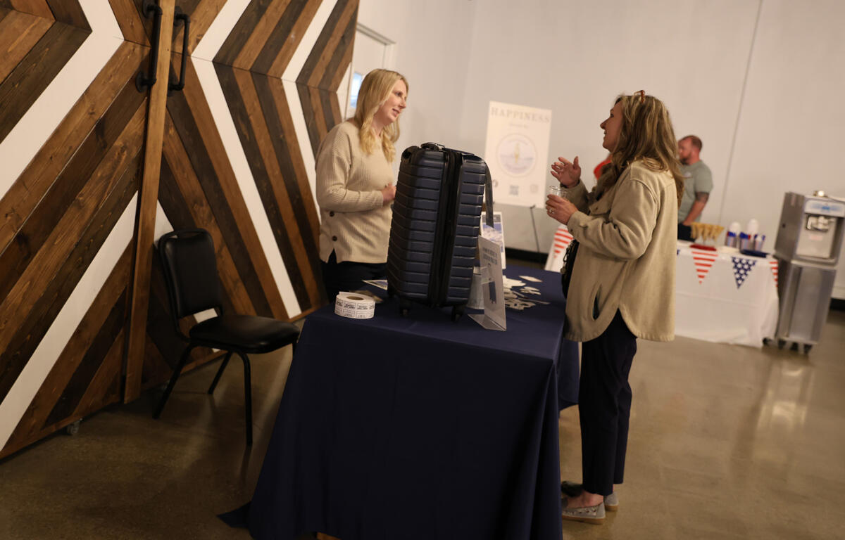 Two women talk at a registration table with a dark suitcase on display in a gallery-like room with wooden geometric wall panels in the background.