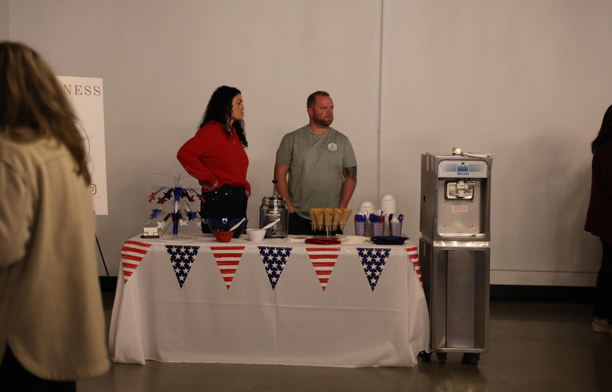 Two people stand behind a dessert table decorated with red, white, and blue bunting, next to a Taylor ice cream machine and cones in cups.