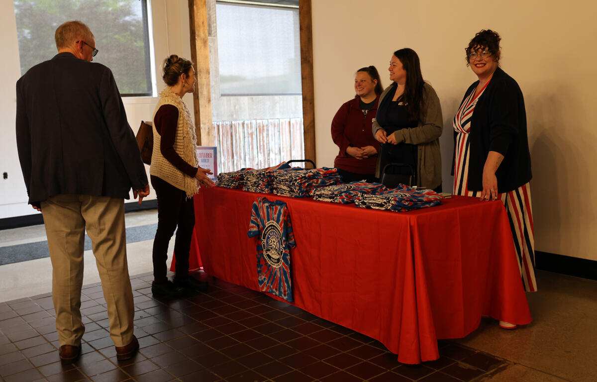 Five people chat around a red-cloth table displaying folded blue and white tie-dye T-shirts at an indoor event.