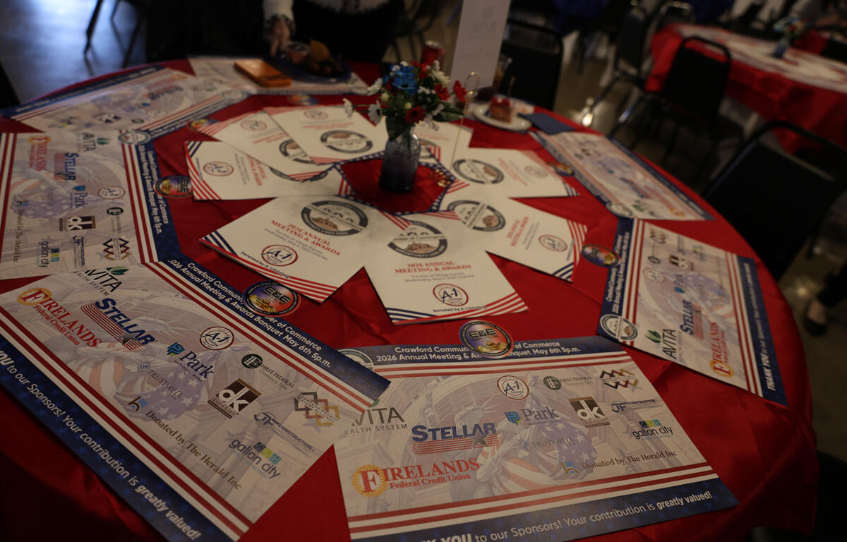 Round table covered with a red cloth displaying event programs, sponsor flyers, and a small floral centerpiece in a blue vase in the middle.