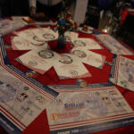 Round table covered with a red cloth displaying event programs, sponsor flyers, and a small floral centerpiece in a blue vase in the middle.