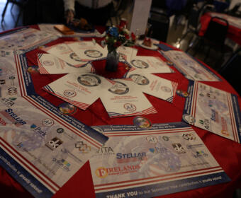 Round table covered with a red cloth displaying event programs, sponsor flyers, and a small floral centerpiece in a blue vase in the middle.