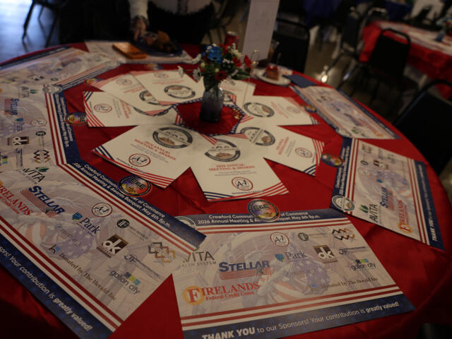 Round table covered with a red cloth displaying event programs, sponsor flyers, and a small floral centerpiece in a blue vase in the middle.