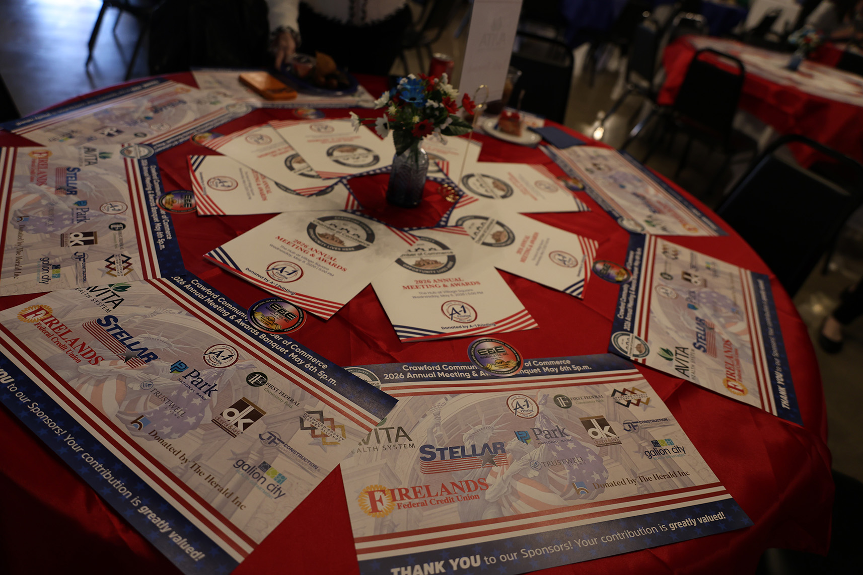 Round table covered with a red cloth displaying event programs, sponsor flyers, and a small floral centerpiece in a blue vase in the middle.