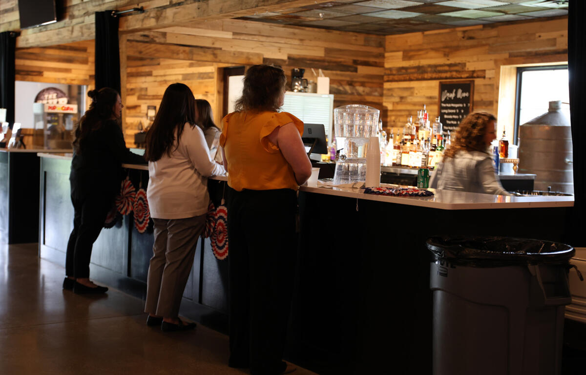 People stand at a curved bar counter in a rustic wood-paneled lounge, waiting to order drinks with bottles and a water dispenser in view behind the bar.