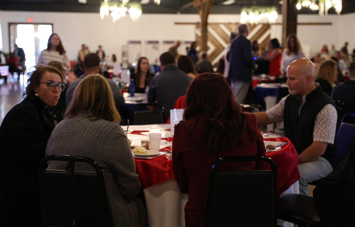 People seated at round tables with red tablecloths in a crowded banquet hall, eating and chatting.