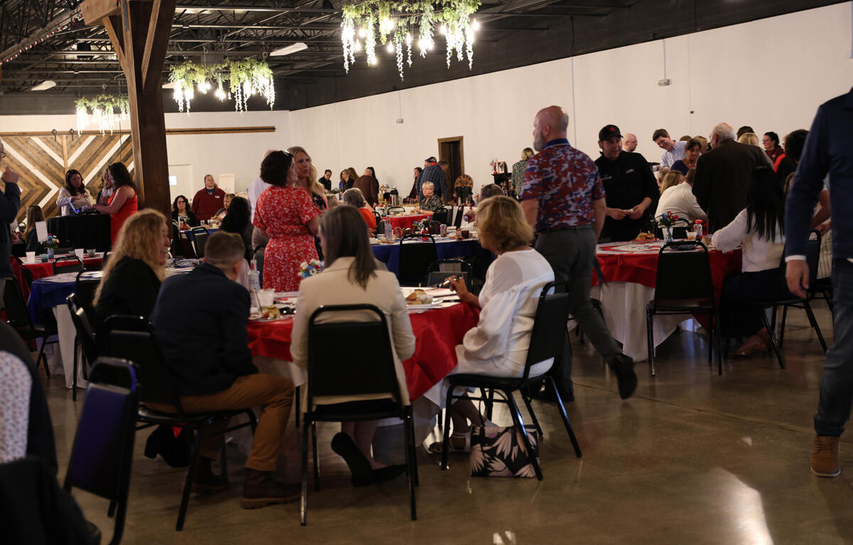 Busy banquet hall with guests seated at round tables covered in red and blue cloths, chatting and dining under hanging greenery and wooden beams.
