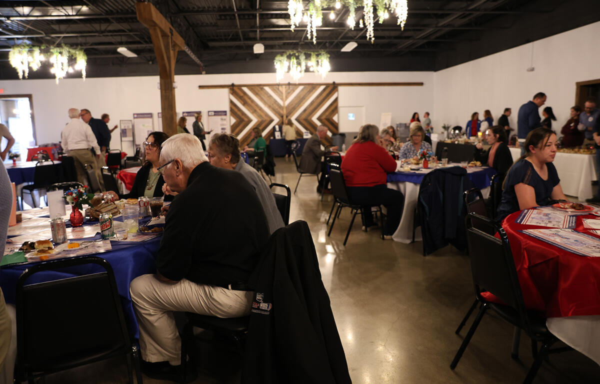 Indoor banquet hall with round tables, people dining, blue tablecloths, and floral centerpieces; wooden chevron wall in back.