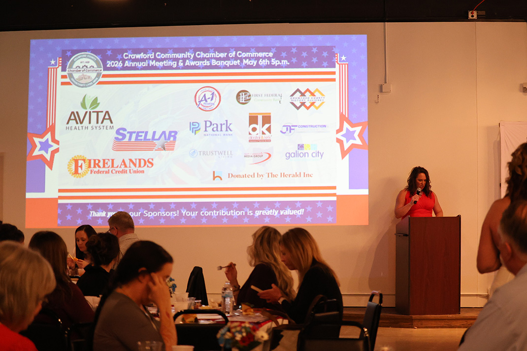 A woman in a red dress speaks at a podium on a stage, with a large screen showing sponsor logos behind her and an audience at banquet tables in the foreground.