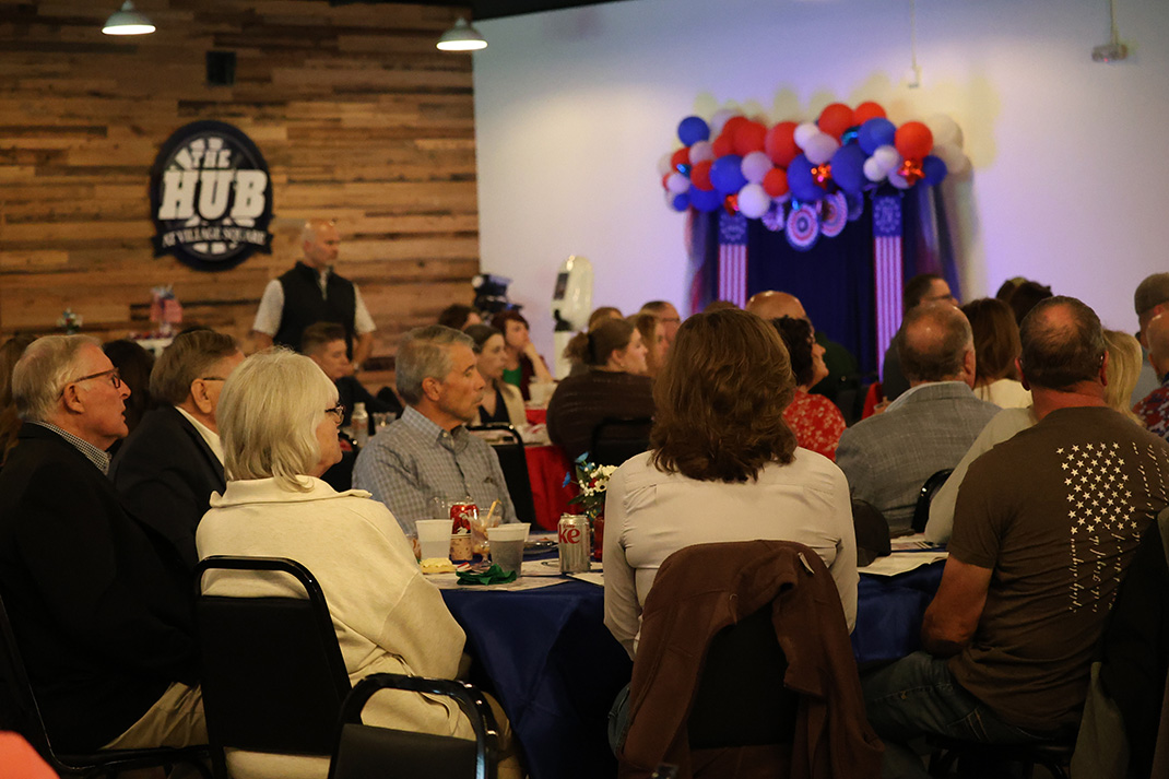 Audience seated at round tables in a hall, facing a stage with a colorful balloon arch and patriotic decor against a wood-paneled wall.