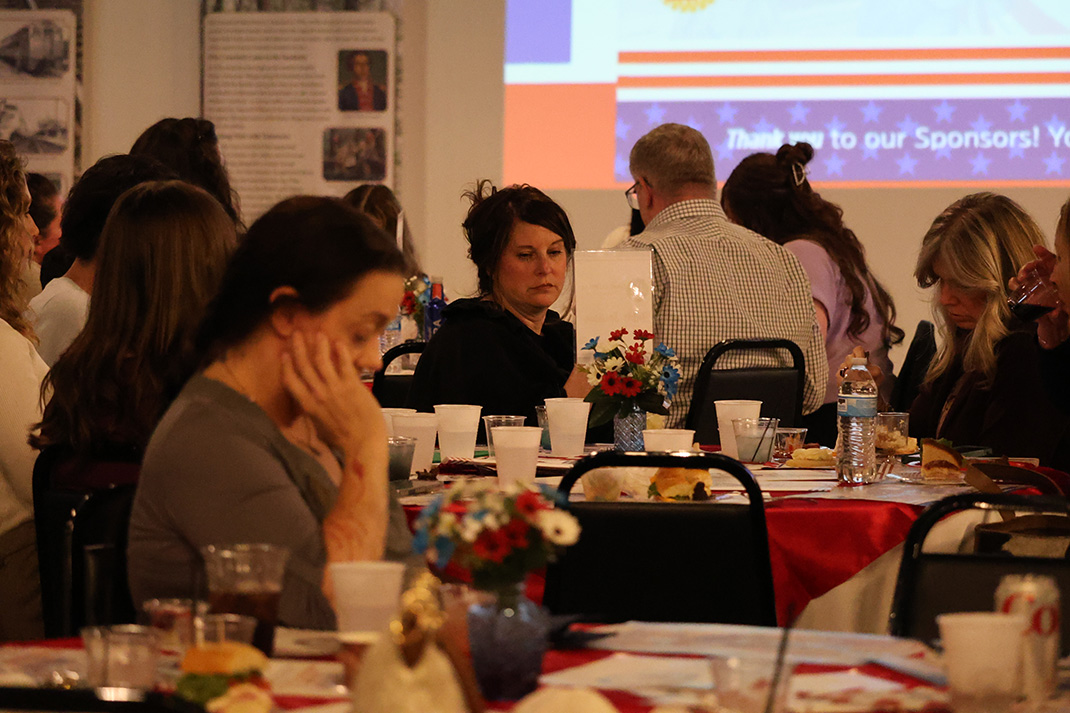 People seated around a long banquet table in a dim room, with plates, drinks, and a patriotic projection on the wall behind them.