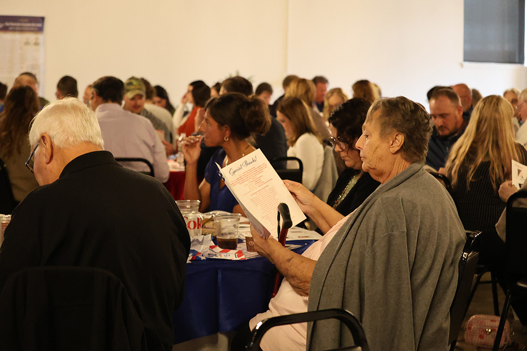 Older woman in a gray sweater reads a menu at a crowded banquet table in a large room