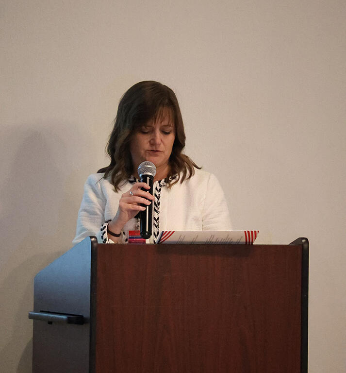 Woman in a white blazer stands at a wooden podium, speaking into a microphone with papers in front of her.
