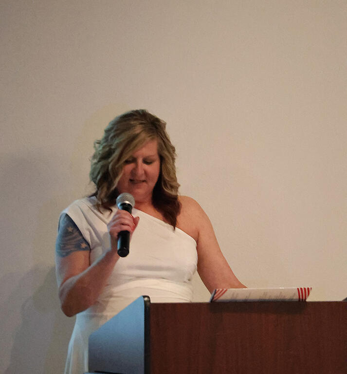 Woman in a white one-shoulder dress stands at a podium, speaking into a handheld microphone in front of a plain wall.