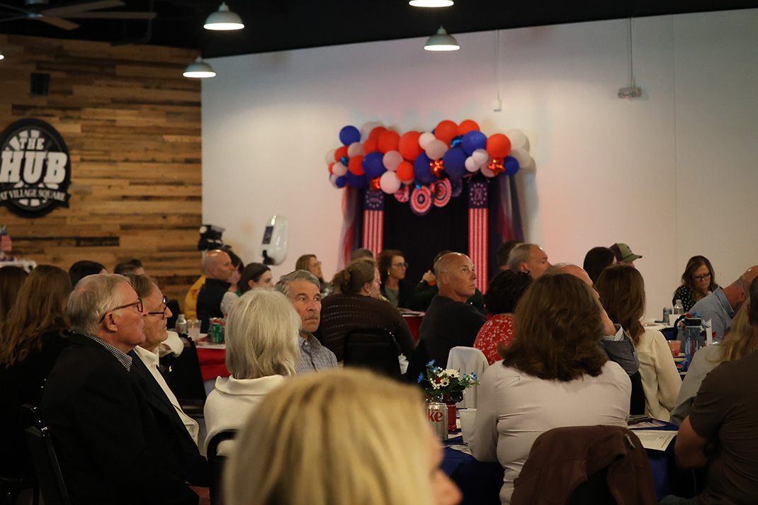 Audience seated at round tables in a hall, facing a stage decorated with red, white, and blue balloons and drapes.
