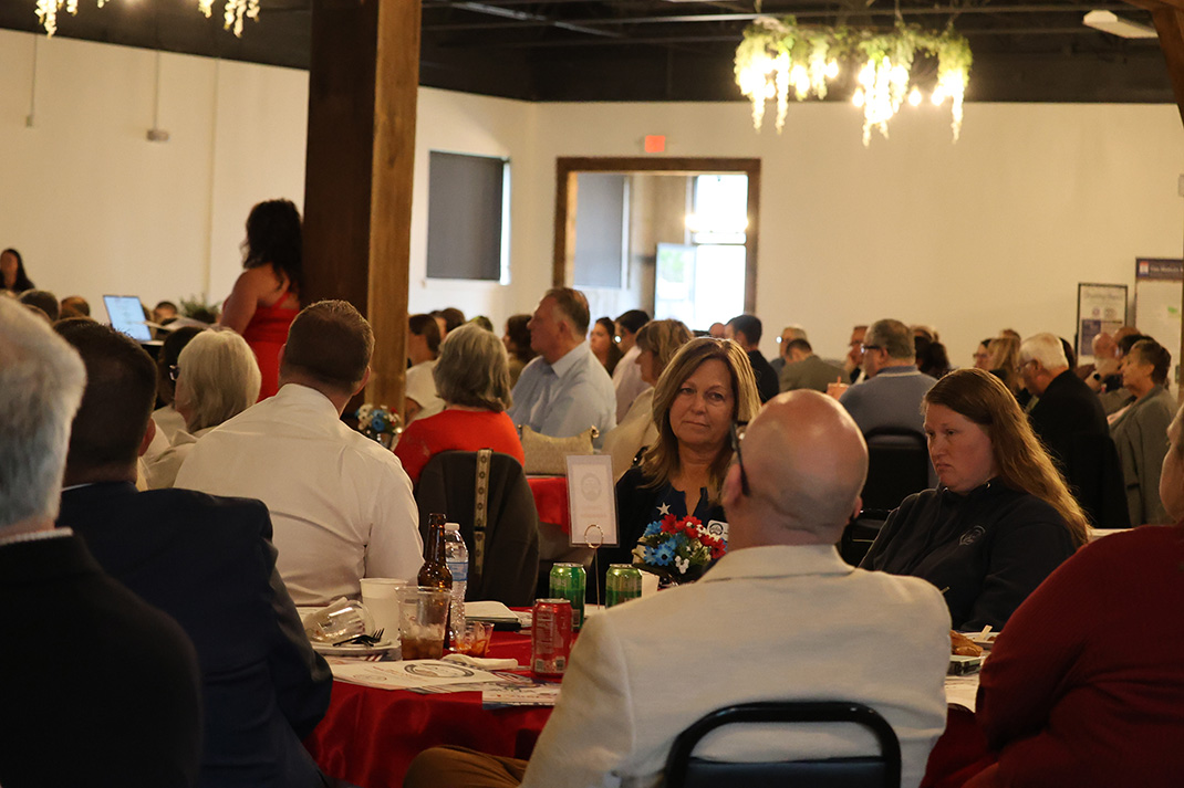 Crowded banquet hall with round tables and people dining at a formal event; a woman in a red dress stands in the back near a pillar.