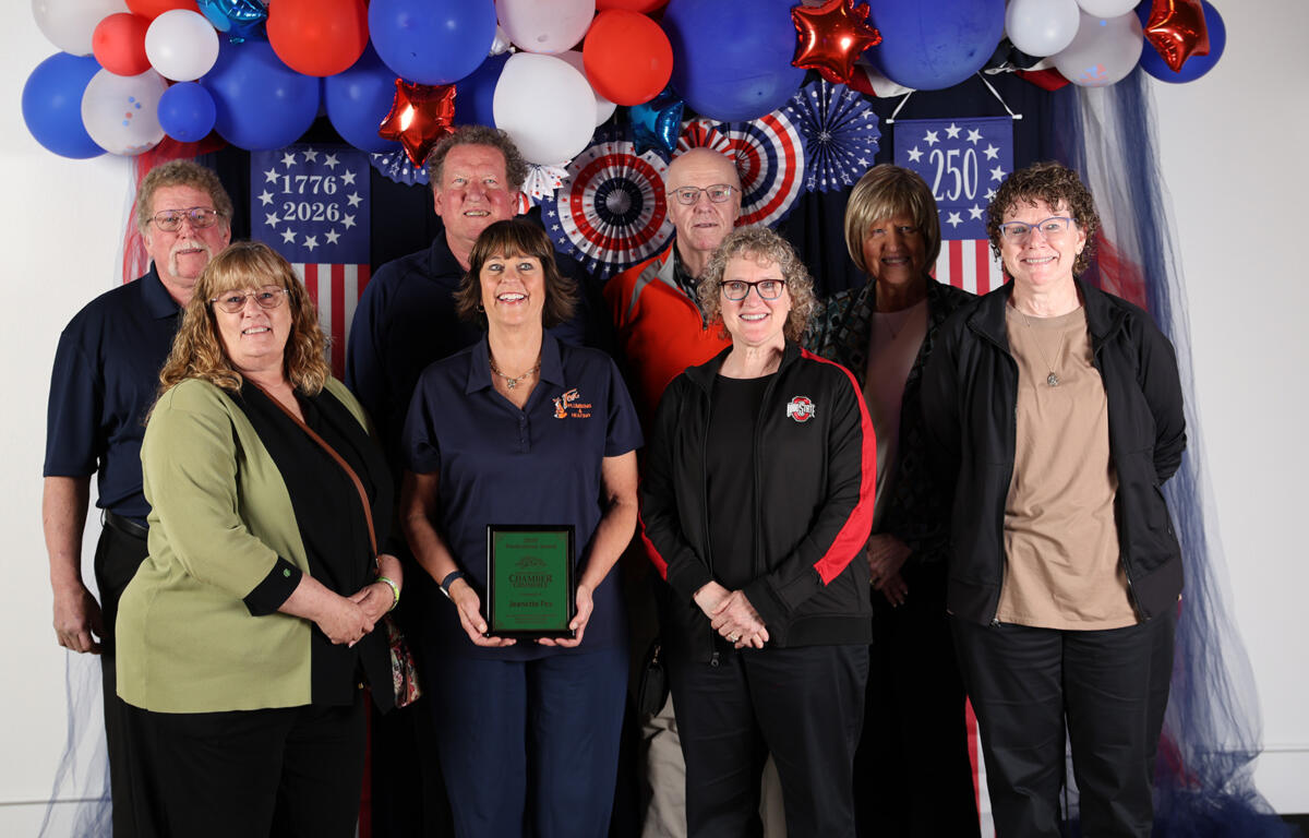 Eight adults pose in a patriotic backdrop with red, white, and blue balloons; a woman in blue scrubs in the center holds a green award plaque.