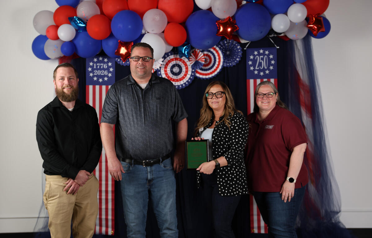 A group of four adults posing for a photo in front of red, white, and blue patriotic decorations; one woman holds a plaque.