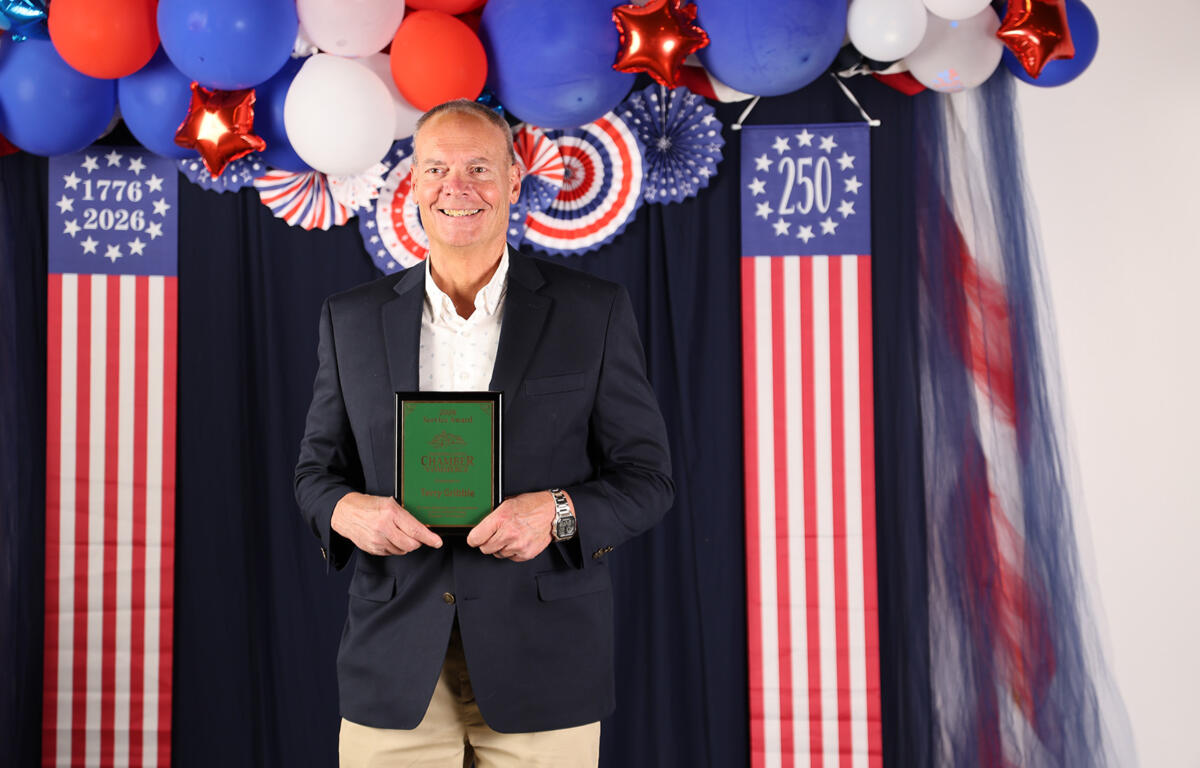 Smiling man in a blazer holds a green award plaque at a patriotic event with red, white, and blue balloons and American flag banners.