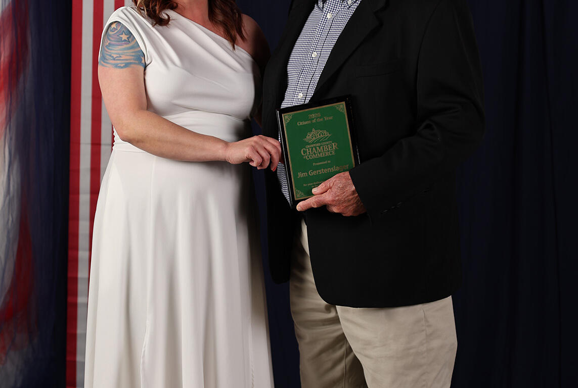 Smiling couple posing at a patriotic event; woman in a white dress beside a man in a blazer who holds a green award plaque, with red, white, and blue decorations behind them.