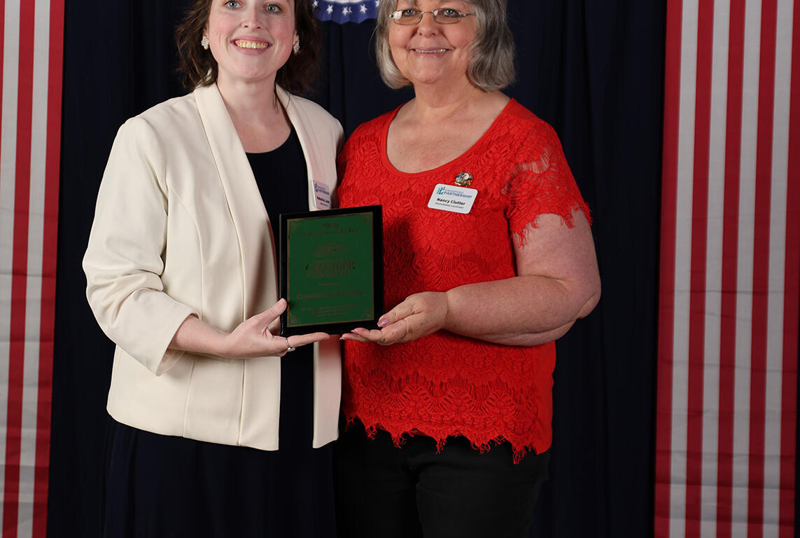 Two women pose for a photo at a patriotic event, holding a green award plaque together in front of red, white, and blue decorations.