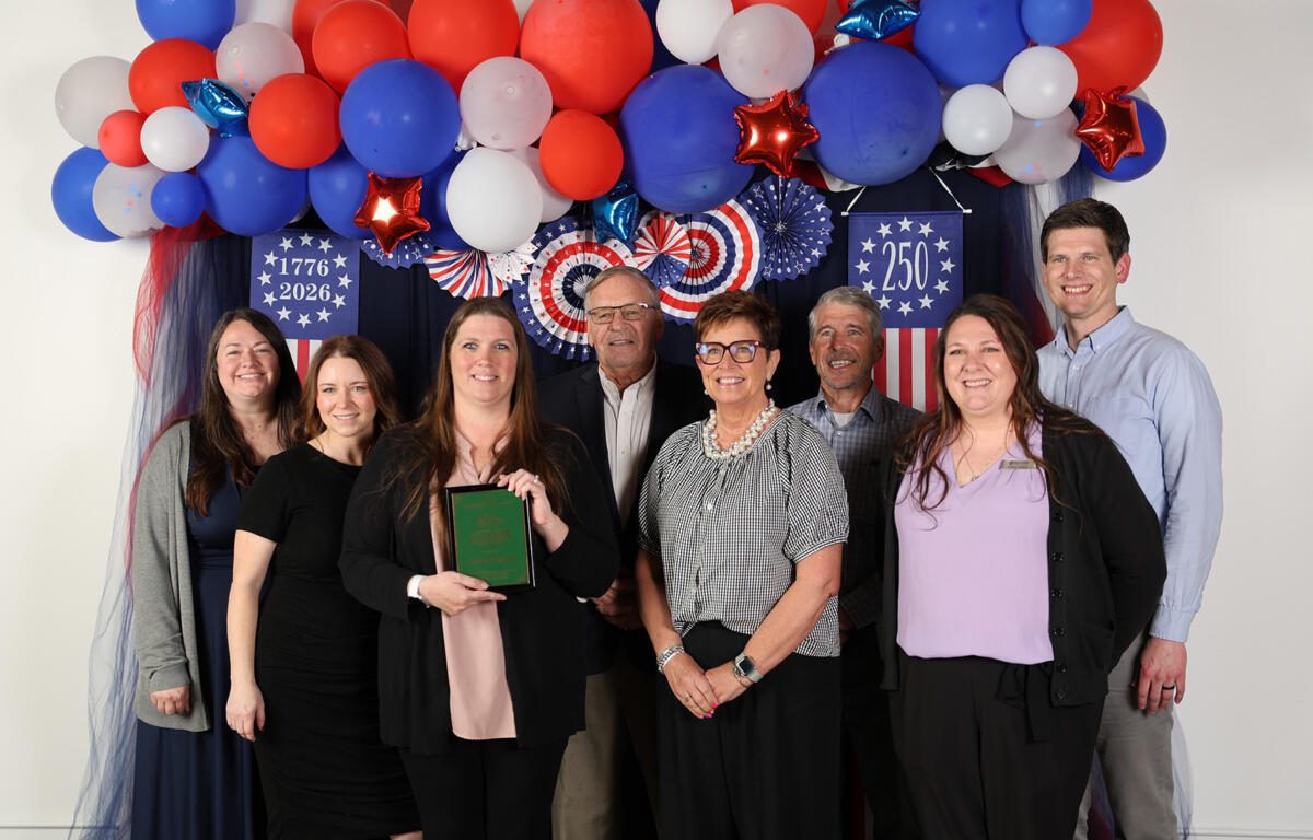Eight people pose together at a patriotic ceremony with red, white, and blue balloons and star banners.