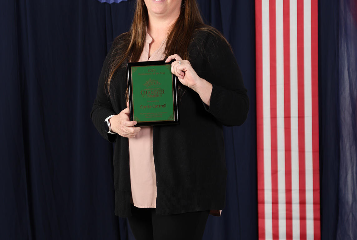 Woman in a black cardigan holding a green award plaque, standing in front of red, white, and blue patriotic decorations and balloons.