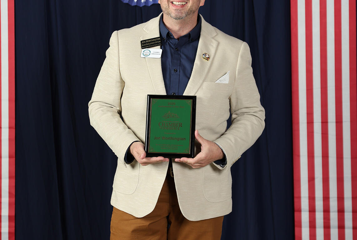 Man in a beige blazer and navy shirt smiles while holding a green award plaque at a patriotic event with red, white, and blue decorations.