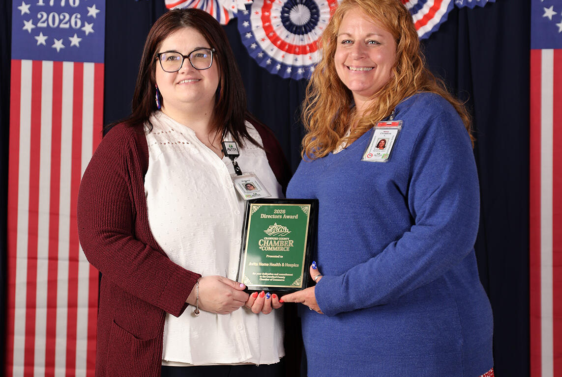 Two women hold a green award plaque together, with red, white, and blue balloons and patriotic decorations behind them.