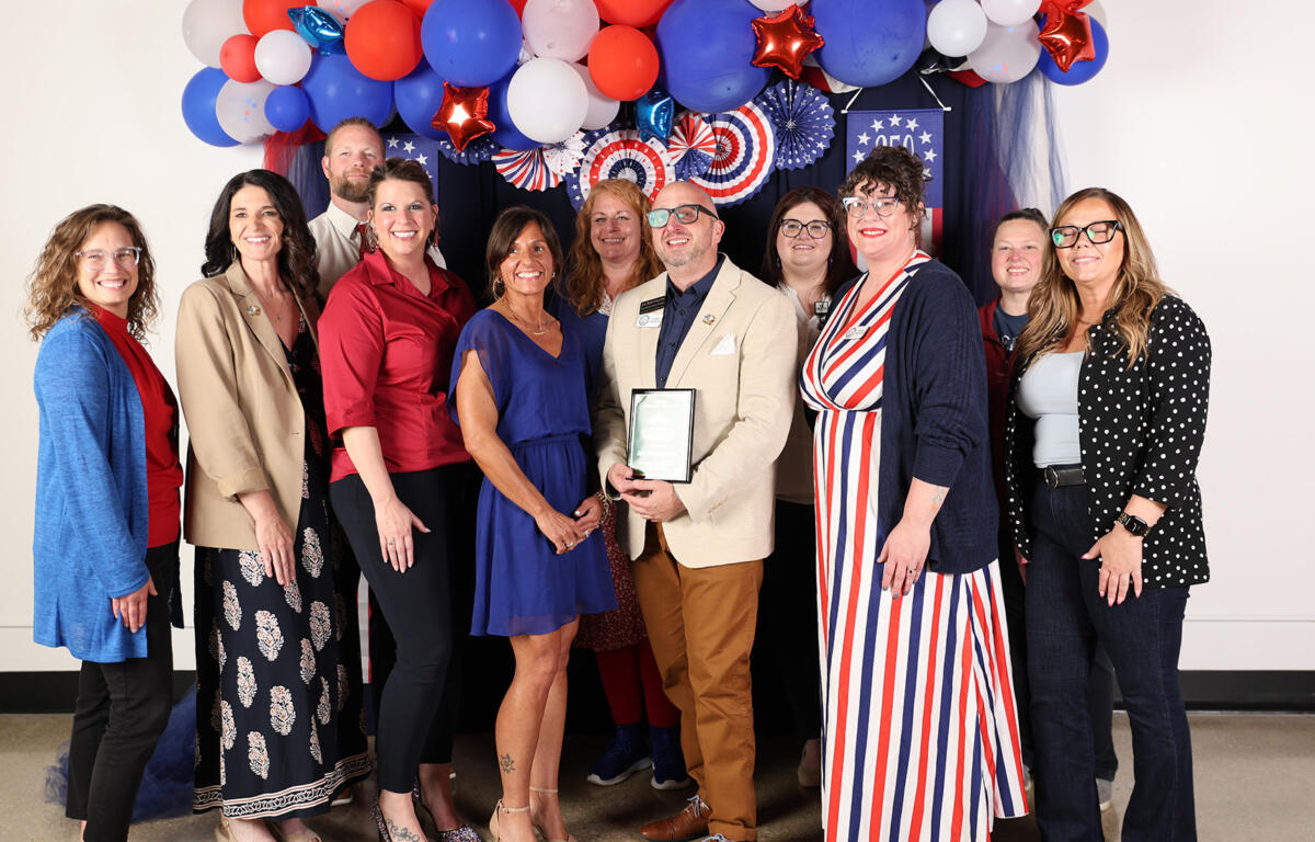 Group of adults posing for a patriotic celebration photo with red, white, and blue balloons overhead; man in a tan blazer holds an award plaque in the center.