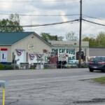 Car dealership and repair shop on a street, with a building displaying 'The Car Guys' sign and a black sedan in the foreground.