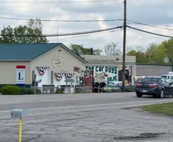Car dealership and repair shop on a street, with a building displaying 'The Car Guys' sign and a black sedan in the foreground.