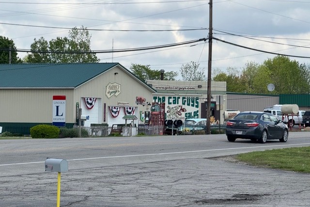 Car dealership and repair shop on a street, with a building displaying 'The Car Guys' sign and a black sedan in the foreground.