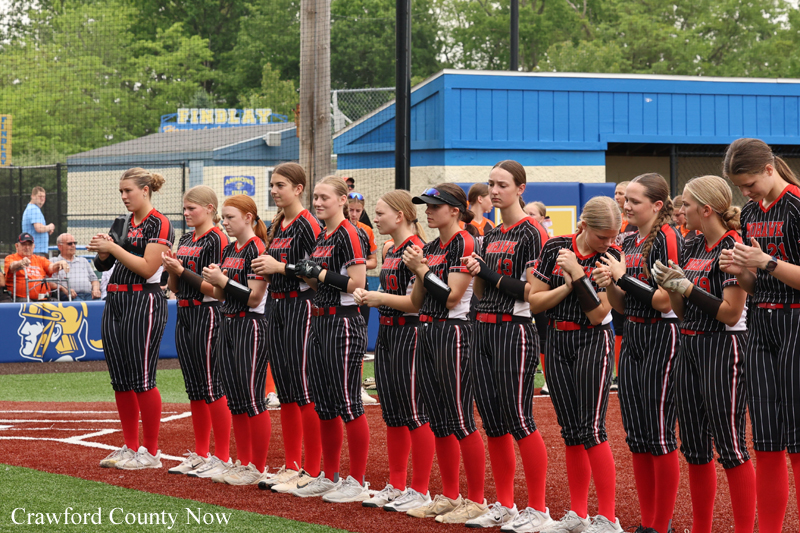 Girls' softball team lines up on the infield, clapping hands as a pre-game ritual, wearing black pinstripe uniforms with red socks.
