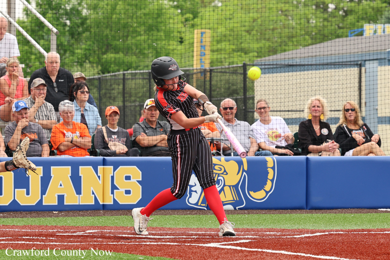 Young female softball player in a black-and-red uniform swings a bat during a game, helmet on, red socks and white cleats, as spectators watch from the stands behind a blue outfield wall.