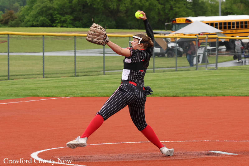 Female softball pitcher in pinstripe uniform delivers a pitch from the mound, right arm raised with ball, glove on left, red socks; fence and school bus in background.