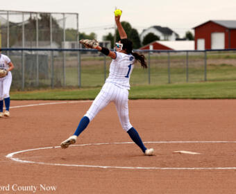 Female softball pitcher in a white uniform and blue socks delivers a pitch from the mound as a teammate watches in the background. The yellow ball is raised in her throwing hand.