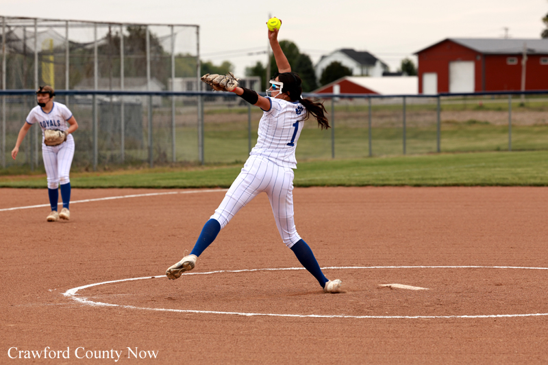 Female softball pitcher in a white uniform and blue socks delivers a pitch from the mound as a teammate watches in the background. The yellow ball is raised in her throwing hand.