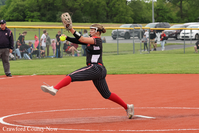 Female softball pitcher in black pinstripe uniform winds up on the mound, releasing a bright ball with spectators in the background.