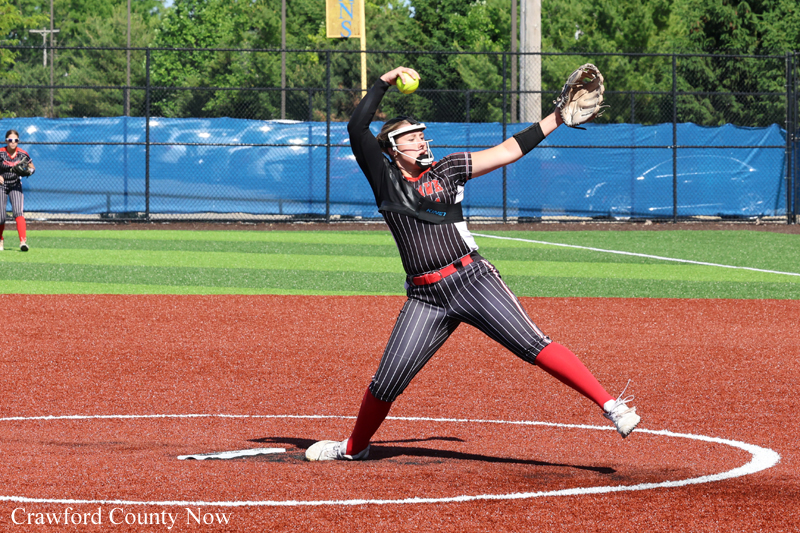 Softball pitcher in a black pinstripe uniform winds up a pitch on a red infield, glove raised and ball ready to throw with a green outfield and blue windscreen in the background.