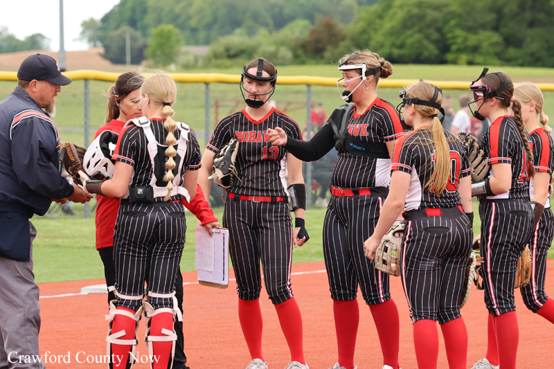 Girls' softball team in pinstripe uniforms with red socks gathers in a huddle on the field, listening to a coach during a timeout.