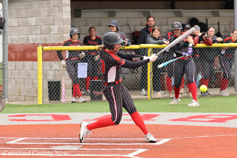 Female softball batter in black-red uniform swings a bat at home plate, crowd watching from the dugout.