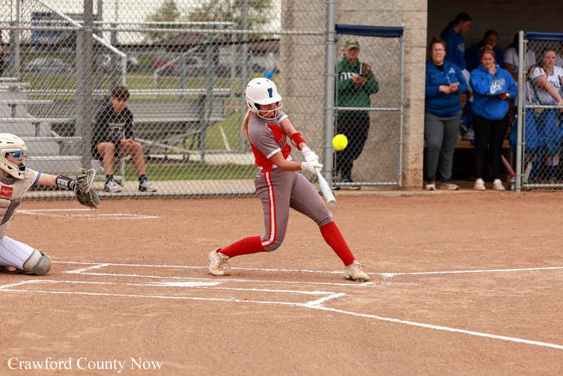 Female softball player in red and gray uniform swings at a pitched ball near home plate, with catcher and bystanders watching in the background.